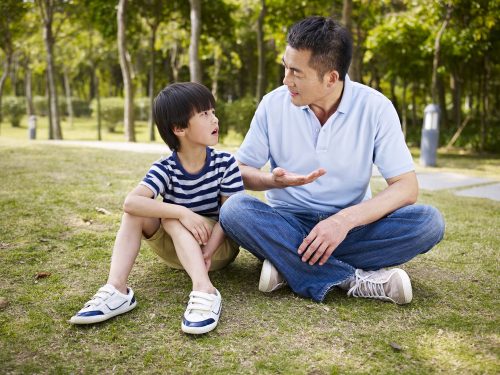 Asian,Father,And,Elementary-age,Son,Sitting,On,Grass,Outdoors,Having
